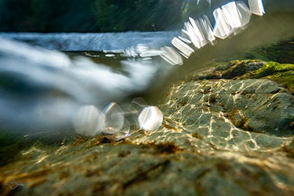 Einblick in die Isar. Am Flußgrund Fels. Darauf reflektiert sich netzförmig das warme, abendliche Sonnenlicht. Die Wasserlinie ist gewellt und glitzert. Bildhintergrund überwasser und im Schatten, Kiestrand und Waldrand.